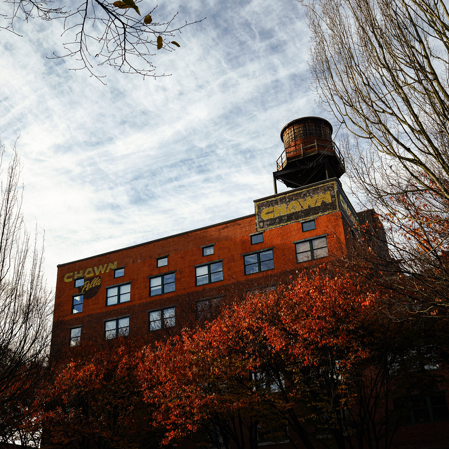 A brick building with a water tower on top is partially obscured by autumn trees under a cloudy sky.
