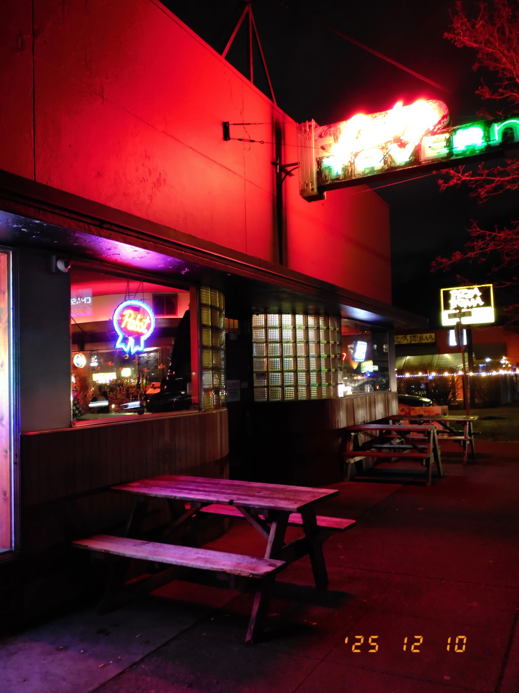 A dimly lit street scene features a bar with neon signs, wooden picnic tables outside, and a warm, inviting glow coming from the windows.