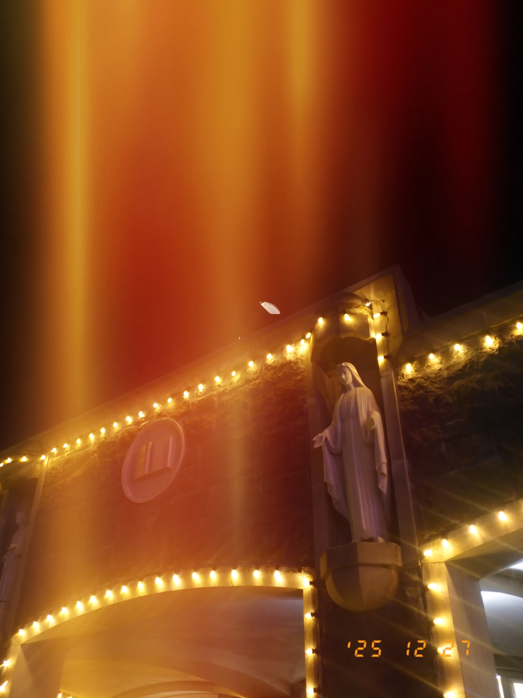 A stone building adorned with string lights features a statue of a religious figure under a dark sky with a glow effect.