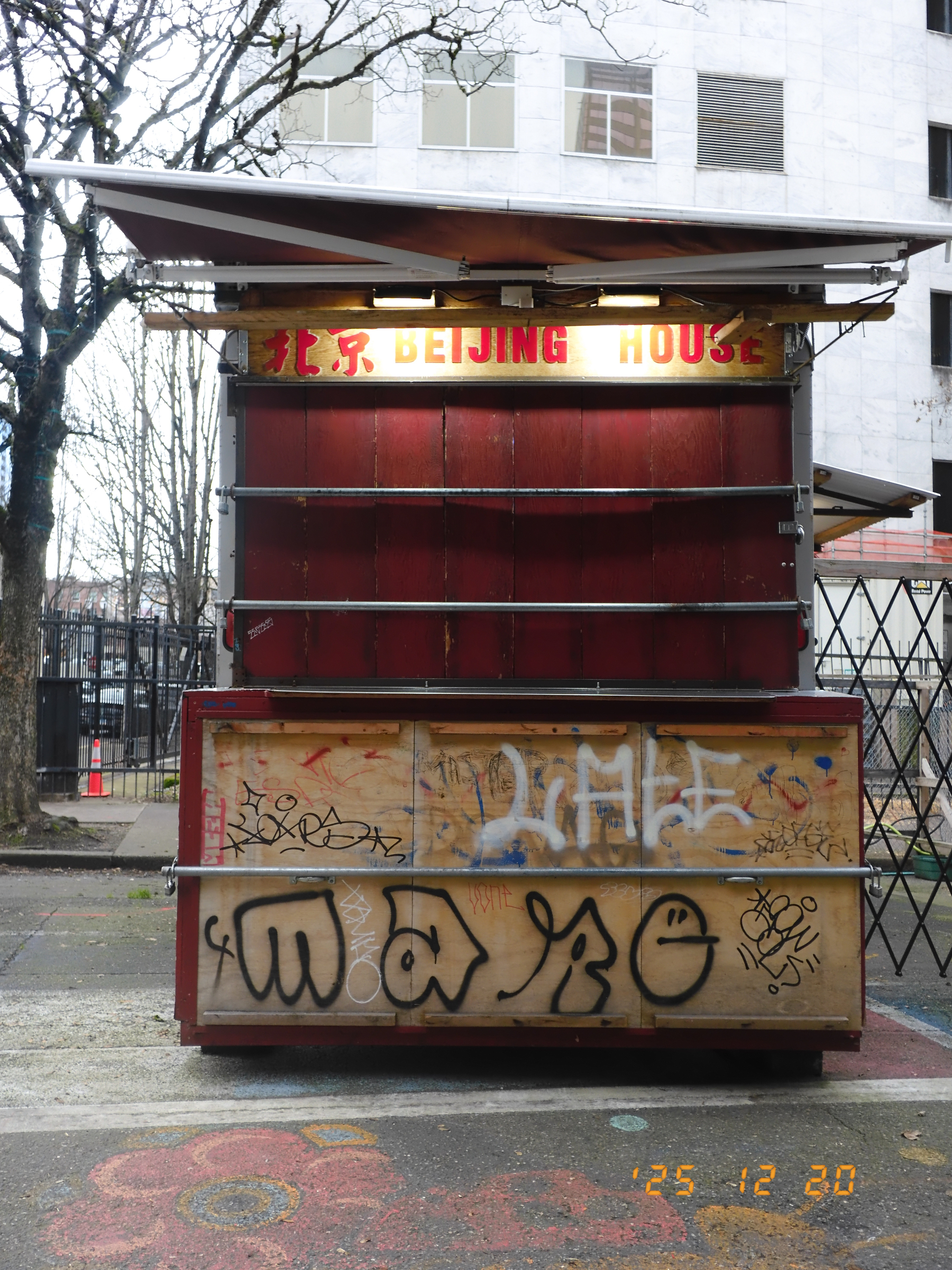 A closed food stand with graffiti on its lower section is set against an urban backdrop.