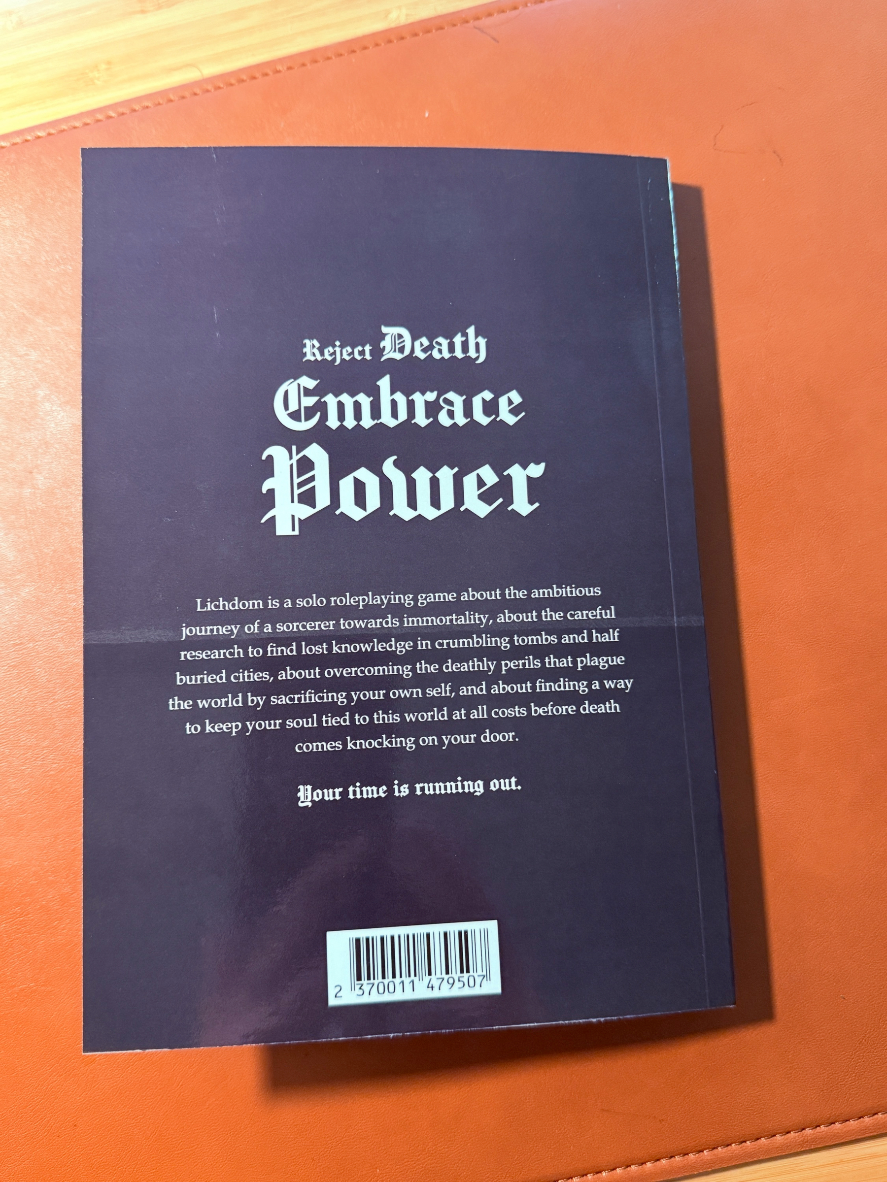 A book with the text “Reject Death, Embrace Power” on an orange surface contains text about a roleplaying game involving a journey toi lichdom and overcoming challenges before death.
