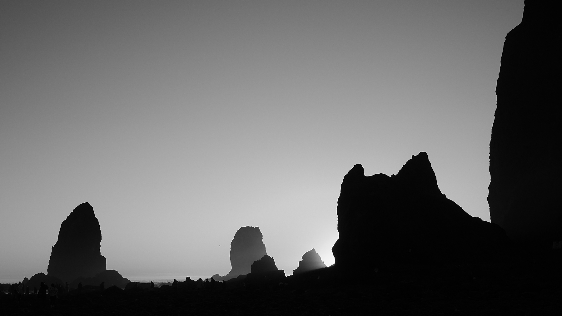 Silhouettes of large sea stacks stand against a clear, sunlit sky along a coastline.