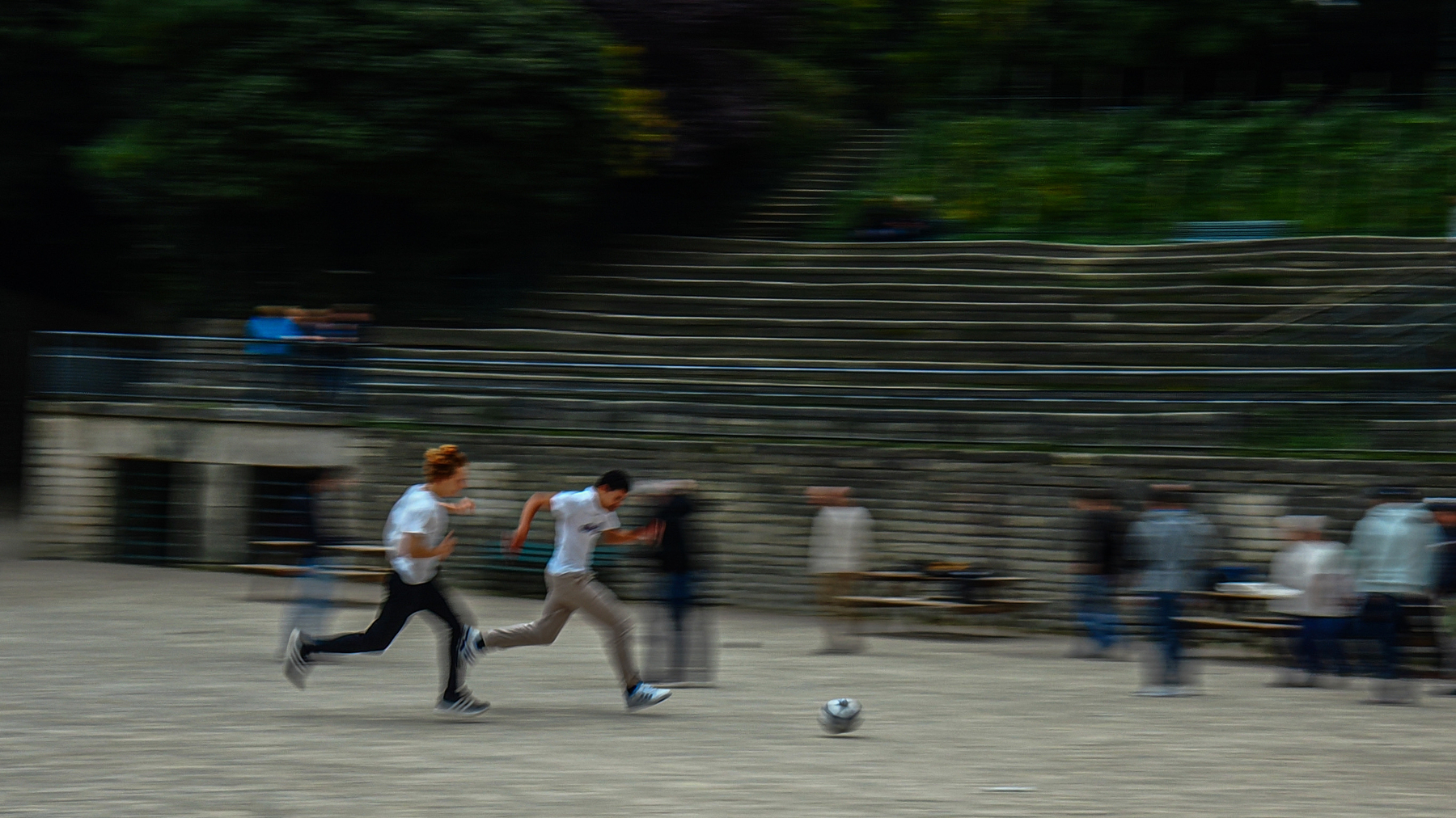 Two people are energetically chasing a soccer ball in what appears to be a public area with blurred motion, while others in the background observe.
