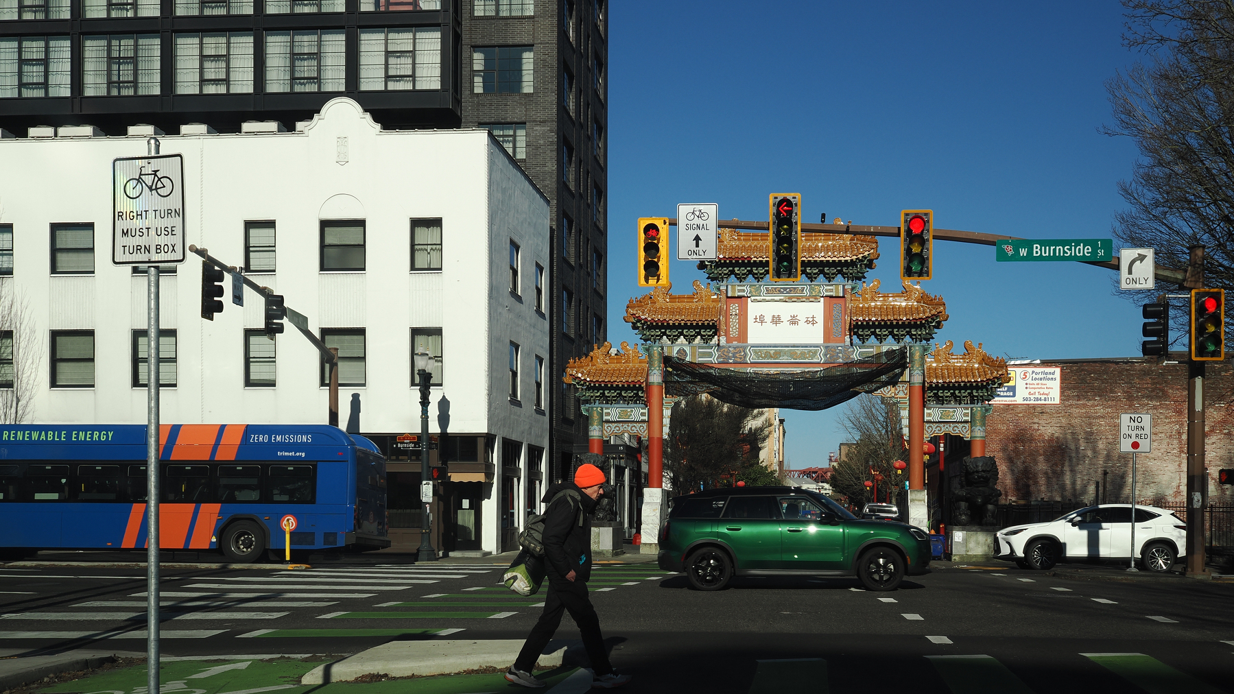 A pedestrian crosses the street near a traditional Chinese gate and various vehicles at an intersection in an urban area.
