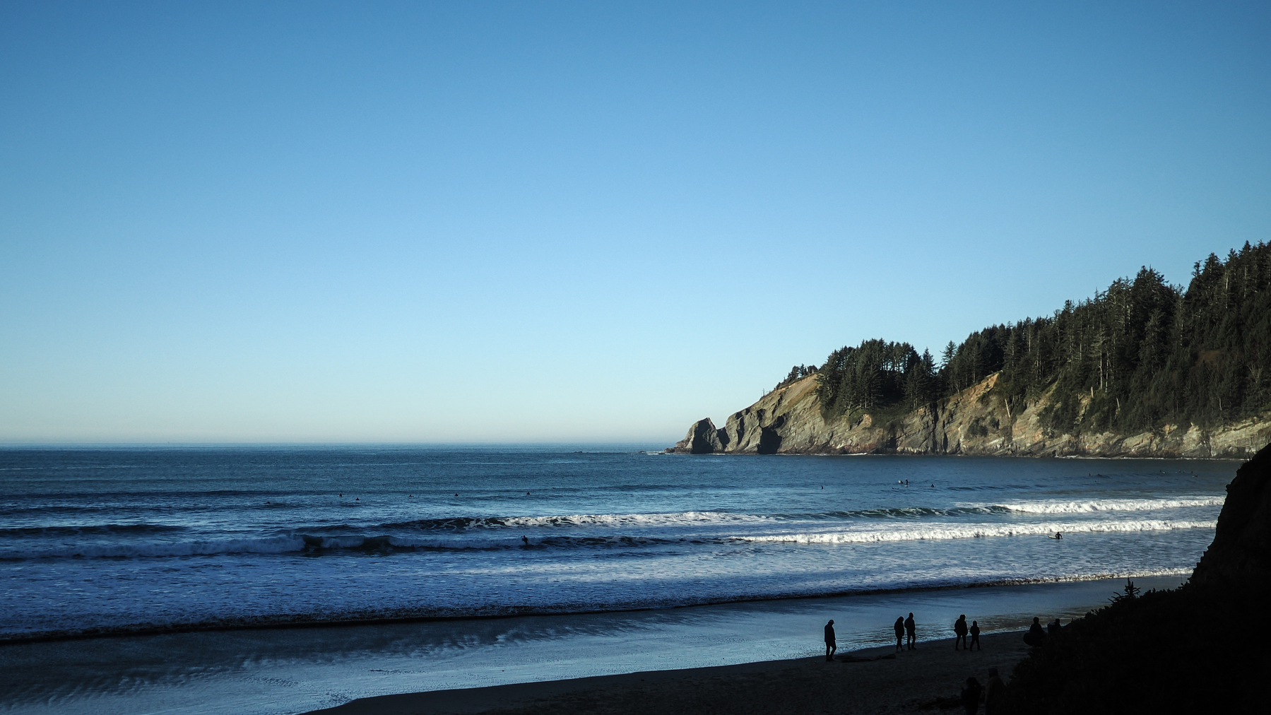 A serene coastal scene features a sandy beach, gentle waves, and a backdrop of forested cliffs.