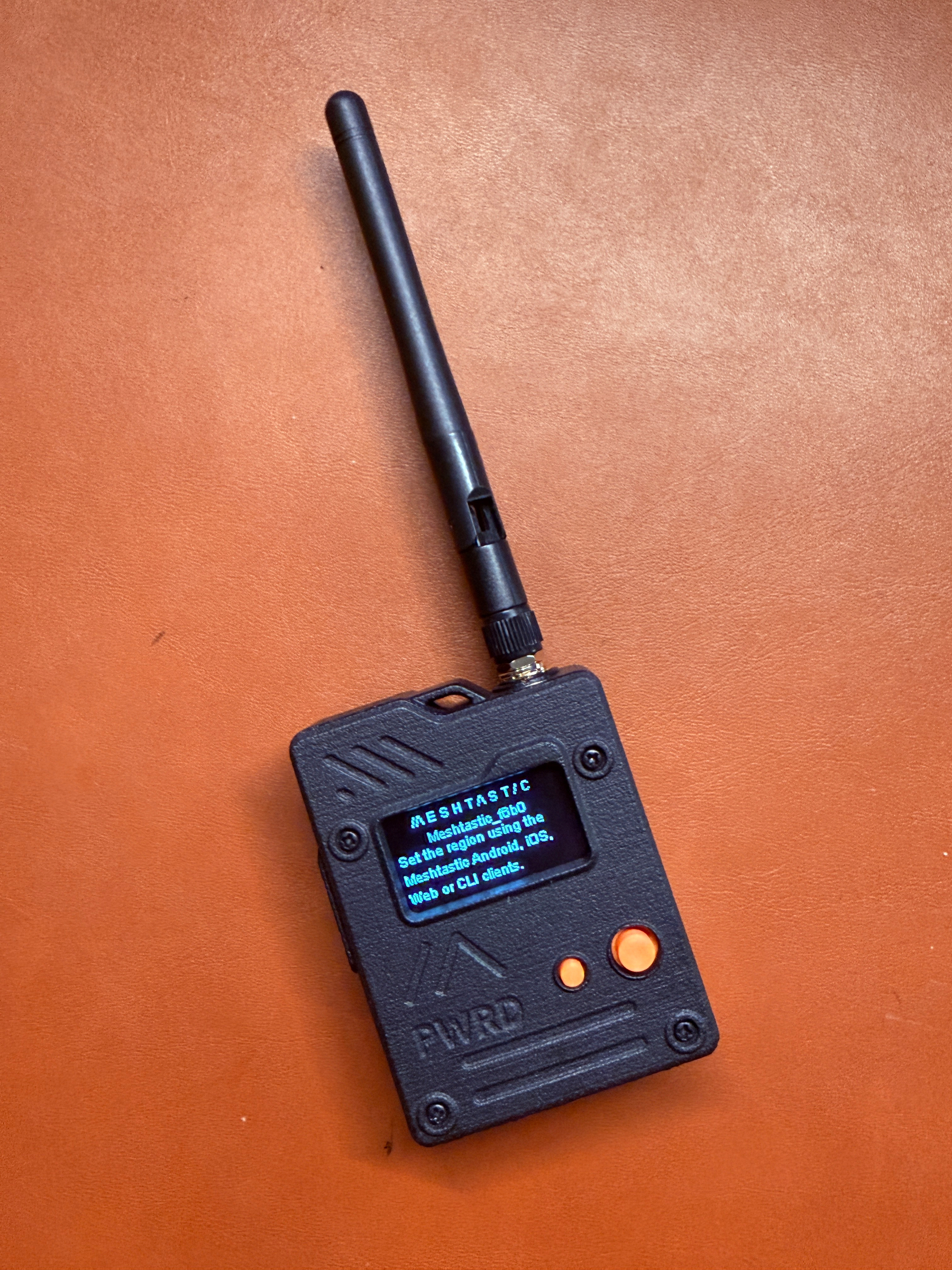 A small black handheld device with an antenna and a screen displaying pixelated text is placed on a brown surface.